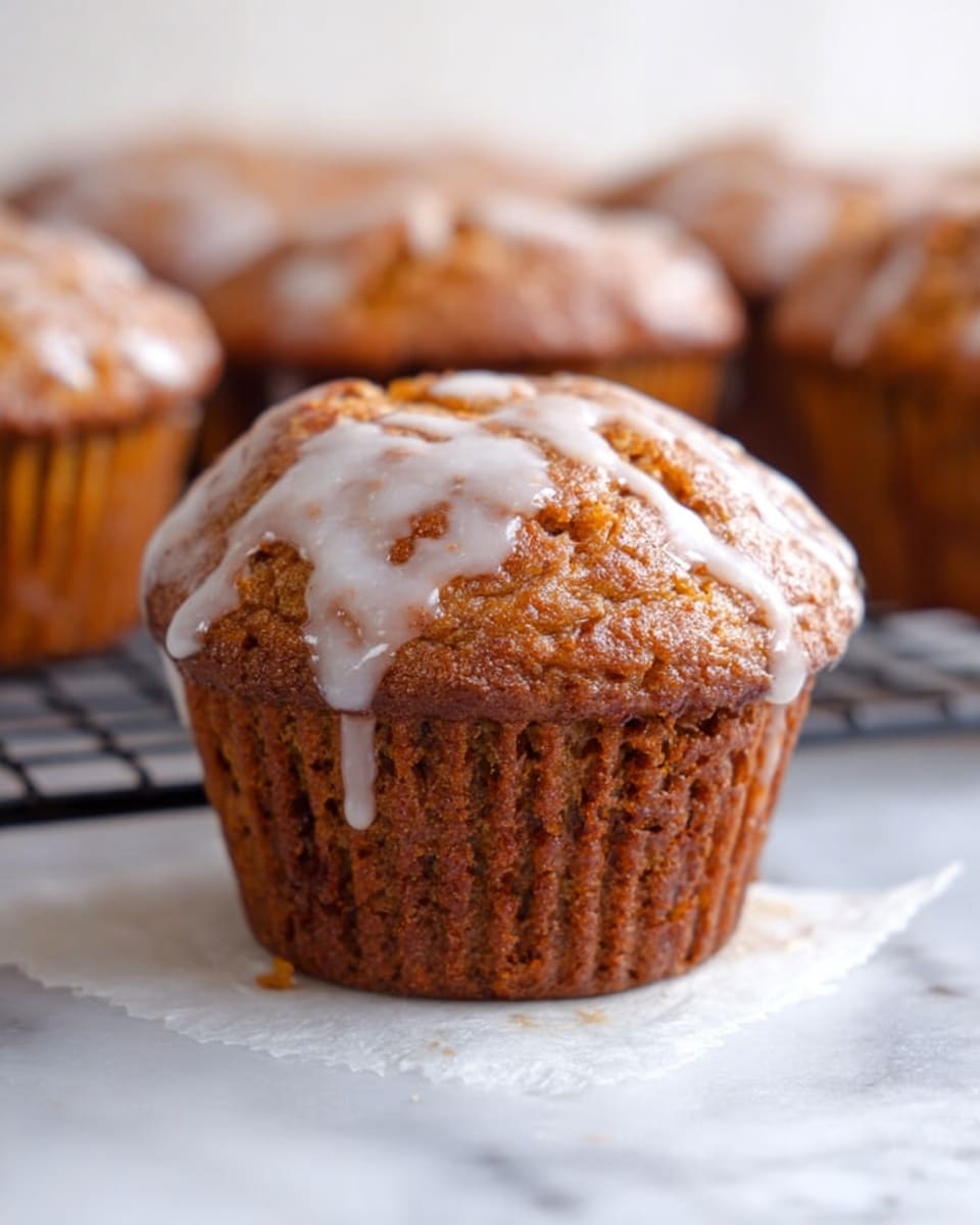 The image shows one brown muffin with a rough, slightly cracked top covered by a thin layer of white icing that drips slightly down the sides. The muffin has a dense texture with a darker brown base and a lighter brown top. Behind it, there are several more muffins, blurred, resting on a white cooling rack. The surface is a white marbled texture with some white paper beneath the muffin in front. Photo taken with an iphone --ar 4:5 --v 7