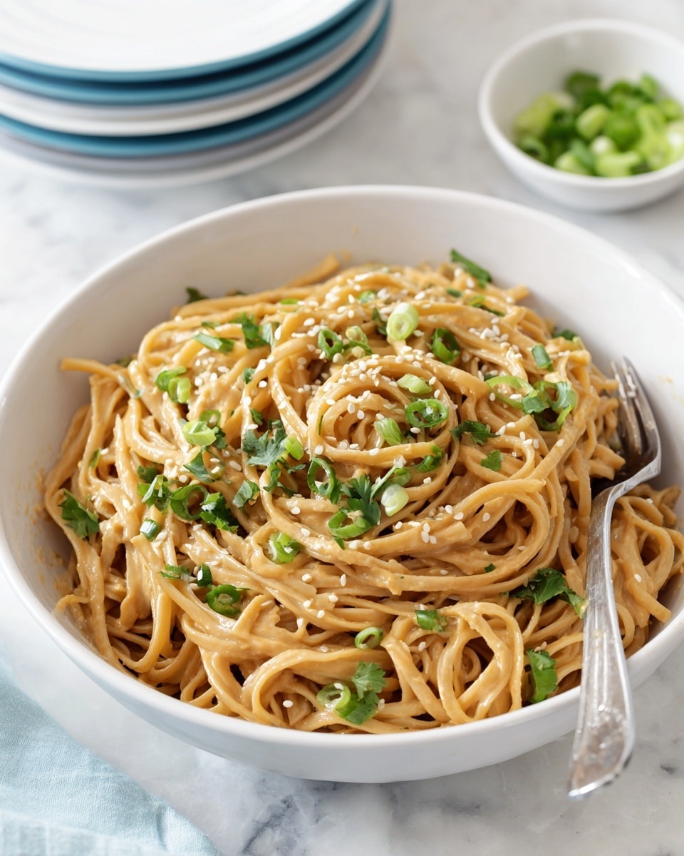 A white bowl filled with creamy peanut noodles, the noodles are light brown and coated evenly with sauce, mixed with chopped green onions and cilantro scattered on top, sprinkled with sesame seeds, a silver serving fork rests inside the bowl on the right side. The bowl sits on a white marbled surface, with a small white bowl of additional sliced green onions partially visible in the background on the top right corner and a stack of white plates with a blue plate on top on the right edge. Photo taken with an iphone --ar 4:5 --v 7