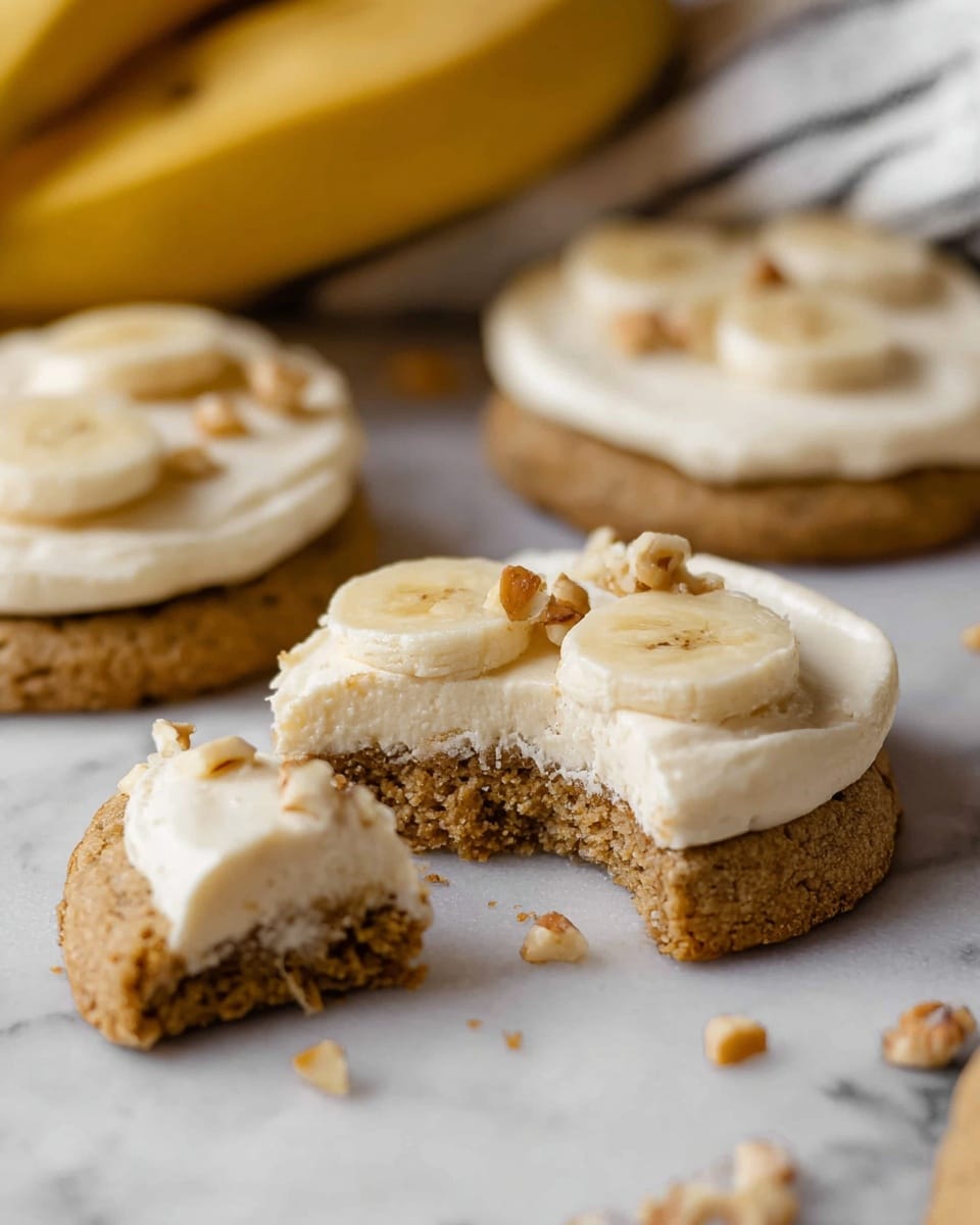 The image shows a close-up of round cookies on a white marbled surface. Each cookie has two layers: a thick, crumbly light brown base and a thick creamy white frosting on top. The frosting is smooth with small banana pieces and light brown nut pieces placed on the surface. One cookie is cut in half, showing the soft texture of the frosting and the dense cookie base. In the background, a bunch of bananas and a white cloth with black stripes are visible but blurred. Photo taken with an iphone --ar 4:5 --v 7