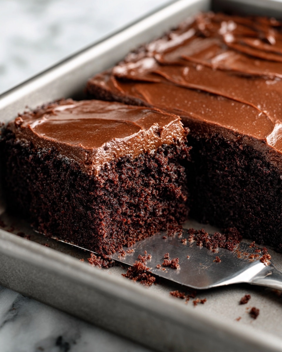 A close-up view of a moist chocolate cake in a silver baking pan, with one slice cut out from the corner. The cake has a thick, smooth top layer of rich, glossy chocolate frosting with a slight shine, while the inside is dark brown, soft, and crumbly with a dense texture. A metal spatula with some chocolate crumbs rests inside the pan under the cut slice. The background is a white marbled texture. photo taken with an iphone --ar 4:5 --v 7