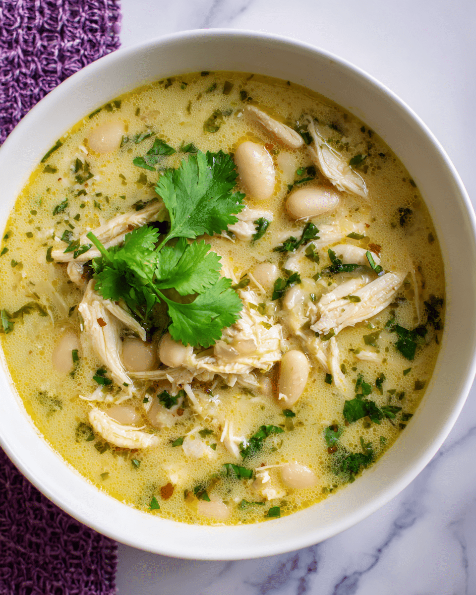 A white bowl filled with a creamy, light yellow soup that has shredded white chicken mixed with white beans and finely chopped green herbs throughout. On top, there are a few fresh, bright green cilantro leaves as garnish, adding color contrast. The soup has a slightly thick texture with visible pieces of chicken and beans. The bowl is placed on a white marbled surface with a purple woven mat underneath. Photo taken with an iphone --ar 4:5 --v 7