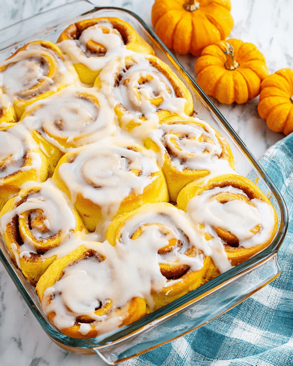 A clear rectangular glass dish filled with nine pumpkin cinnamon rolls arranged in three rows, each roll golden yellow with a soft, fluffy texture showing swirled orange-brown cinnamon filling inside. The rolls are generously covered with a thick, creamy white icing that drapes over the spirals and pools slightly between the rolls. The dish is placed on a white marbled surface with three small orange pumpkins and a blue checkered fabric napkin in the background, adding a cozy autumn feel. photo taken with an iphone --ar 4:5 --v 7