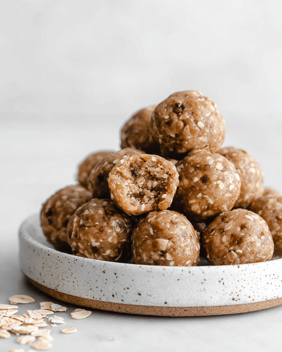 A pile of small round energy balls is stacked on a white speckled plate placed on a white marbled surface. The energy balls have a light brown color with visible oats embedded throughout their slightly rough texture. One energy ball is bitten, showing a dense, chewy inside filled with bits of oats and darker specks, possibly chocolate or nuts. The arrangement is simple and natural with a few scattered oat flakes on the surface near the plate. photo taken with an iphone --ar 4:5 --v 7