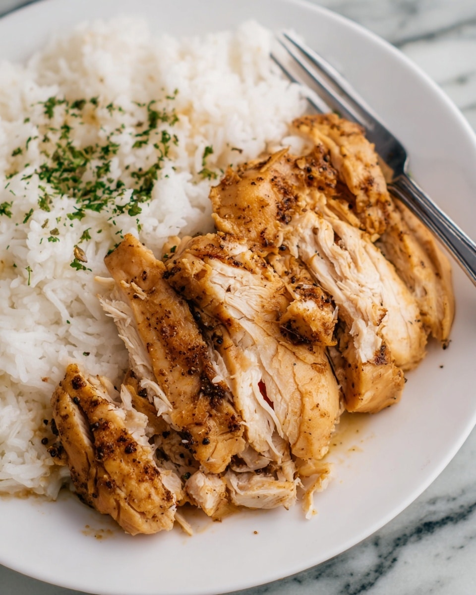 The image shows a close-up of a white plate with two layers of food. The first layer is white rice with some small green herbs sprinkled on top, covering the upper part of the plate. The second layer is several pieces of cooked chicken placed in front of the rice, with a golden brown color and visible black pepper and seasoning on its surface. The chicken has a juicy and slightly shiny texture, with one piece partially shredded to show its tender inside in light cream color. A silver fork is placed on the right side of the plate, partly under the chicken. The plate is set on a white marbled textured surface. Photo taken with an iphone --ar 4:5 --v 7