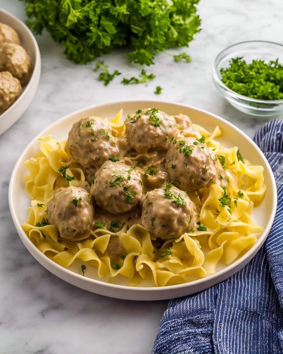 On a round white plate set on a white marbled surface, there is a serving of creamy pasta and meatballs. The bottom layer shows wide, flat, yellow egg noodles coated with a smooth, light brown cream sauce. On top, there are seven medium-sized meatballs with a soft, beige exterior speckled lightly with green herbs. Small bits of chopped green parsley garnish both the pasta and meatballs, adding a fresh touch. Nearby, there is a bunch of fresh parsley, a blue-striped cloth, and a glass bowl filled with more chopped parsley. Photo taken with an iphone --ar 4:5 --v 7