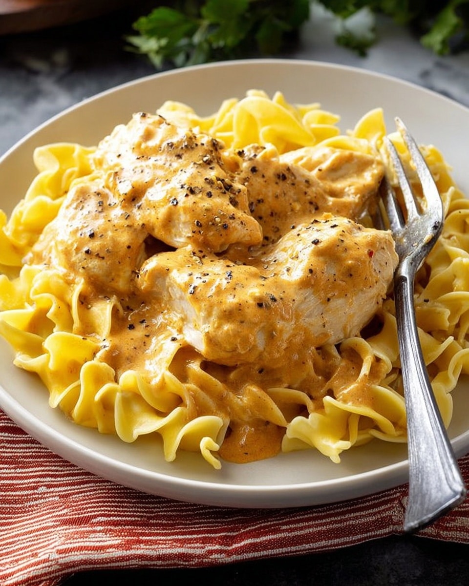 The dish shows a white plate filled with curly yellow egg noodles forming the bottom layer, appearing soft and slightly twisted. On top, there are three pieces of chicken covered in a thick, creamy light orange sauce, which also spreads over parts of the noodles. Black pepper is sprinkled over the sauce and chicken, giving texture and contrast. A silver fork rests on the right side of the plate. The plate is placed on a white marbled surface with a red-striped cloth underneath, and some green leaves appear blurred in the background. Photo taken with an iphone --ar 4:5 --v 7
