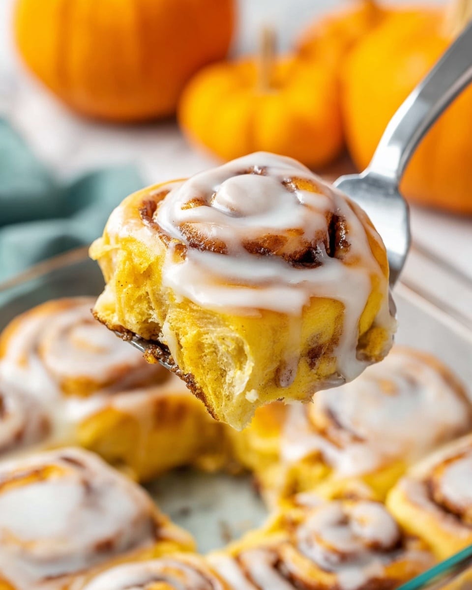 A close-up view of a single pumpkin cinnamon roll being lifted with a metal spatula, showing one thick layer of soft, golden-yellow dough swirled with cinnamon brown and topped with a smooth, white glaze that slightly drips down the edges. The roll’s texture looks moist and fluffy with visible cinnamon streaks inside. In the background, more cinnamon rolls with the same layers and glaze are in a clear glass baking dish, set on a white marbled surface. There are small orange pumpkins blurred in the background, adding an autumn feel. Photo taken with an iphone --ar 4:5 --v 7