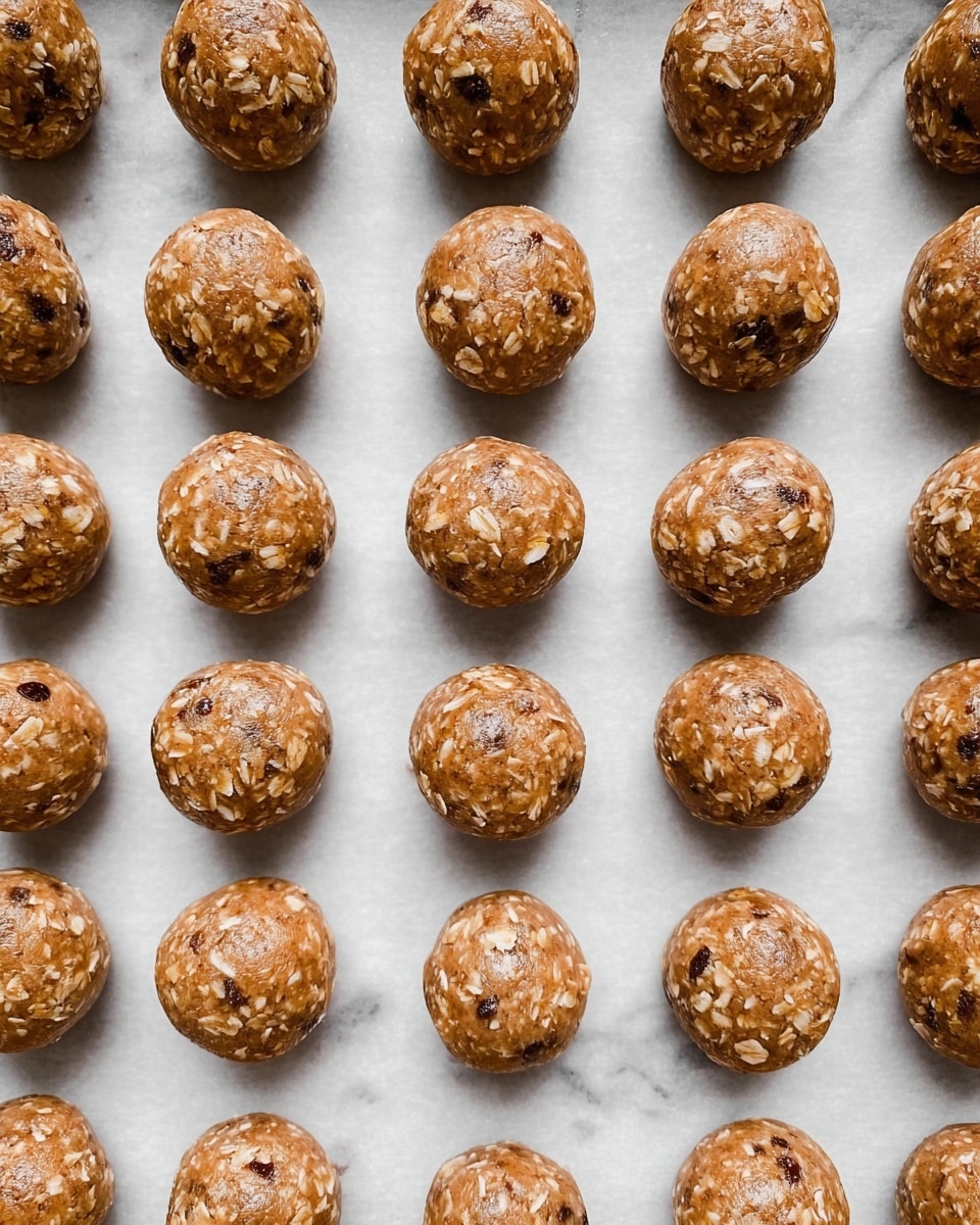 The image shows a single layer of round cookie dough balls arranged in neat rows on a white parchment paper on a baking tray. The dough balls have a light brown color with visible white oat flakes and darker small chocolate or nut pieces mixed inside. Each ball has a smooth but slightly textured surface, giving them a rustic, homemade look. The parchment paper and tray fill the entire background, which is a white marbled texture. photo taken with an iphone --ar 4:5 --v 7