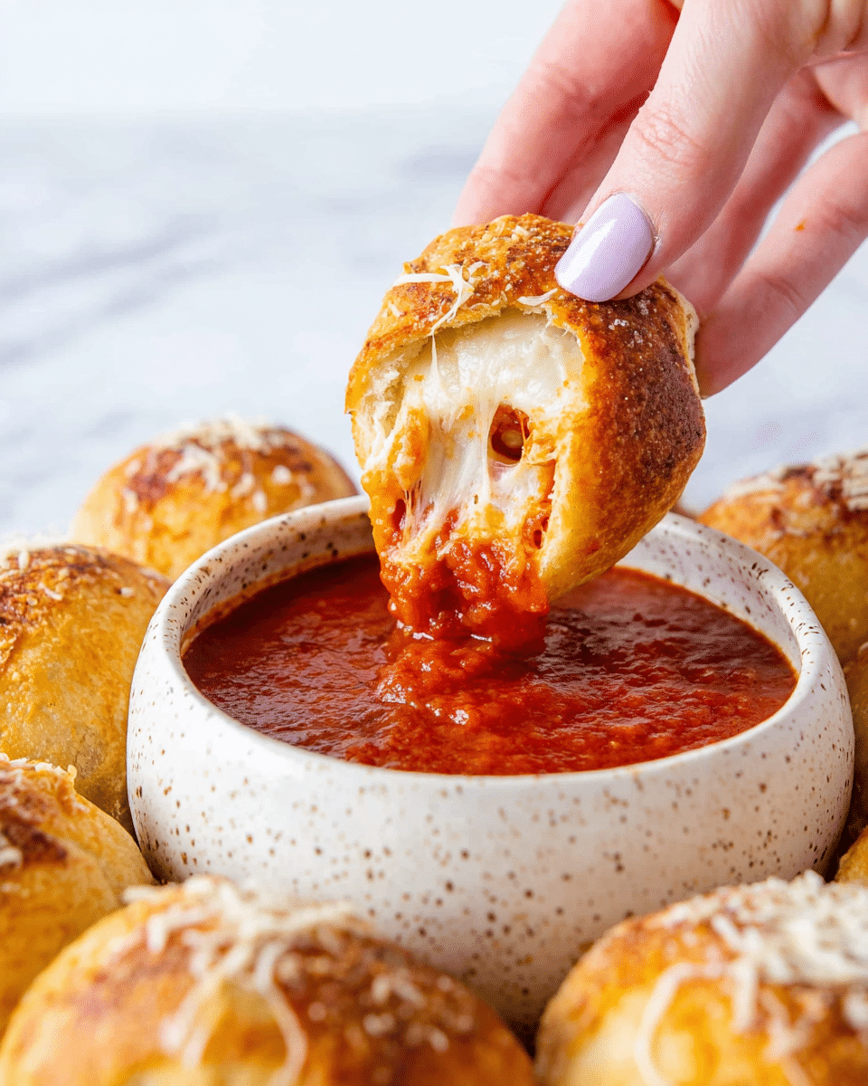A woman's hand with light pink nail polish is holding a stuffed bread piece dipped into thick red marinara sauce in a white speckled bowl. The bread piece has an outer golden-brown crust with a soft inside filled with melted white cheese and small pieces of pepperoni, showing a gooey stretchy texture. Around the bowl, there are more golden brown stuffed bread rolls sprinkled lightly with cheese on top, all placed on a white marbled surface. Photo taken with an iphone --ar 4:5 --v 7