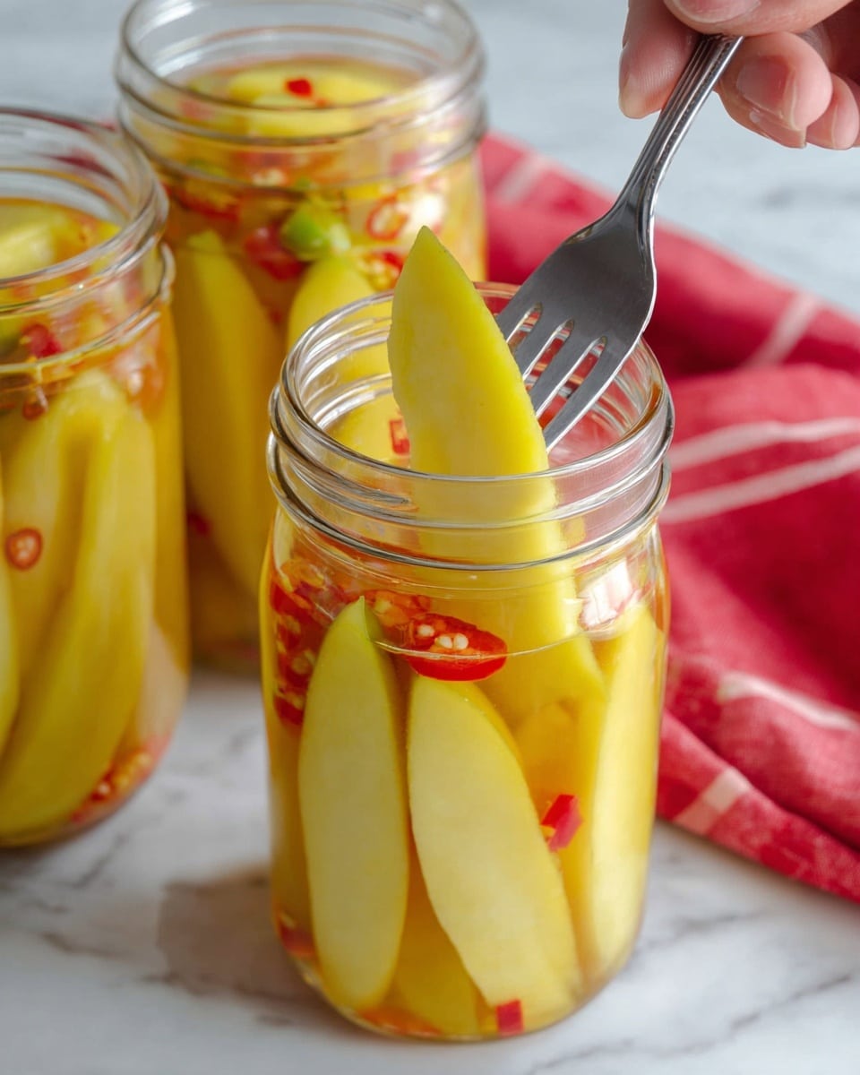 The image shows a clear glass jar filled with long, pale yellow pickled mango slices standing vertically inside, immersed in a clear brine with small red chili slices floating near the top. Behind this jar, two more similar jars are visible, also filled with mango slices in brine. The jars are placed on a white marbled texture, with a metal fork with a wooden handle on the right side and a white cloth with red stripes folded on the left side. photo taken with an iphone --ar 4:5 --v 7