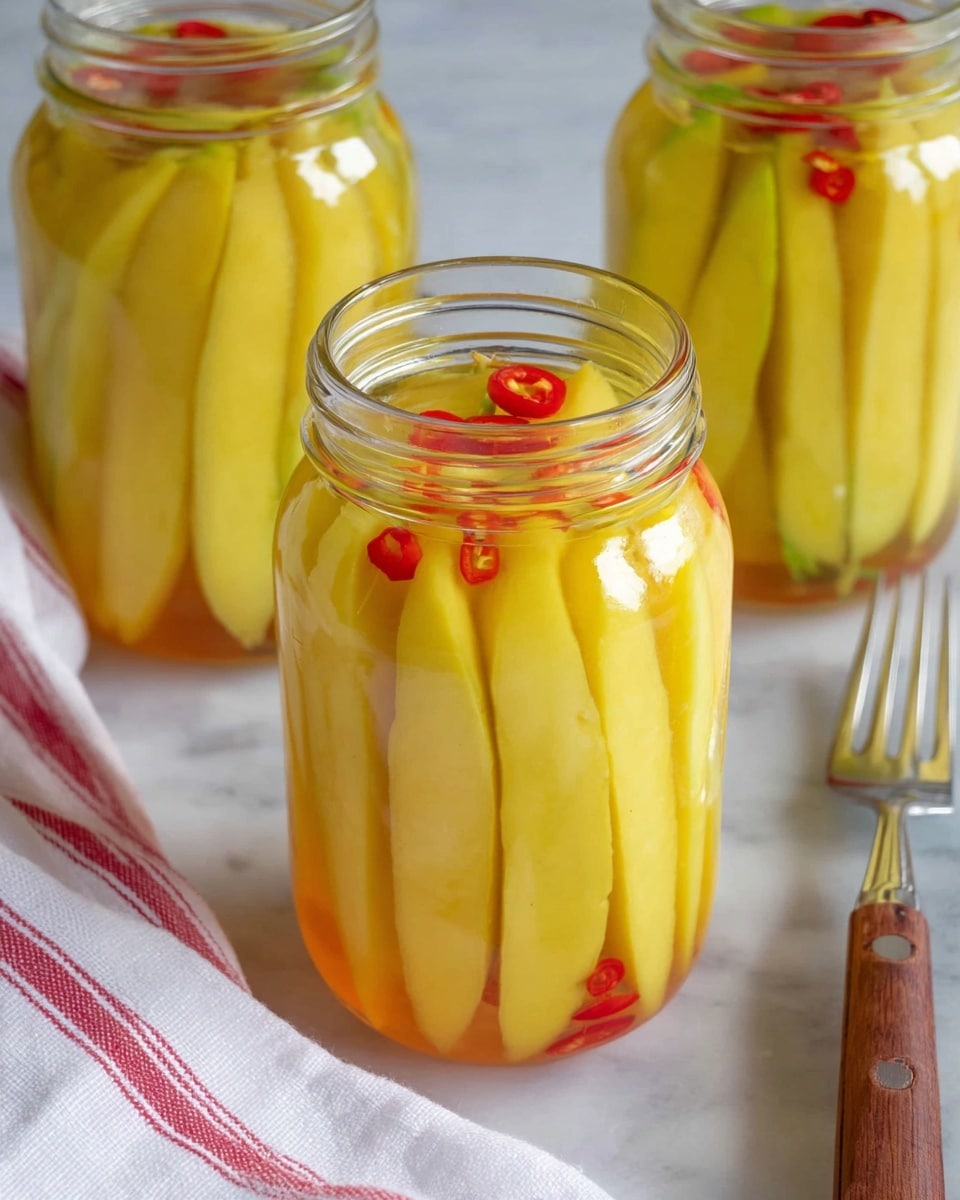 A close-up of a clear glass jar filled with long, pale yellow slices of pickled mangoes, mixed with small round red chili slices evenly spread throughout the jar. A silver fork is held by a woman's hand gently lifting one thick, smooth-textured mango slice from the jar's opening. Behind the jar, two more similar jars filled with the same pickled mango slices are visible, all placed on a white marbled surface with a red and white cloth next to them. The pickled mangoes have a slightly translucent, glossy look with a moist texture. Photo taken with an iphone --ar 4:5 --v 7