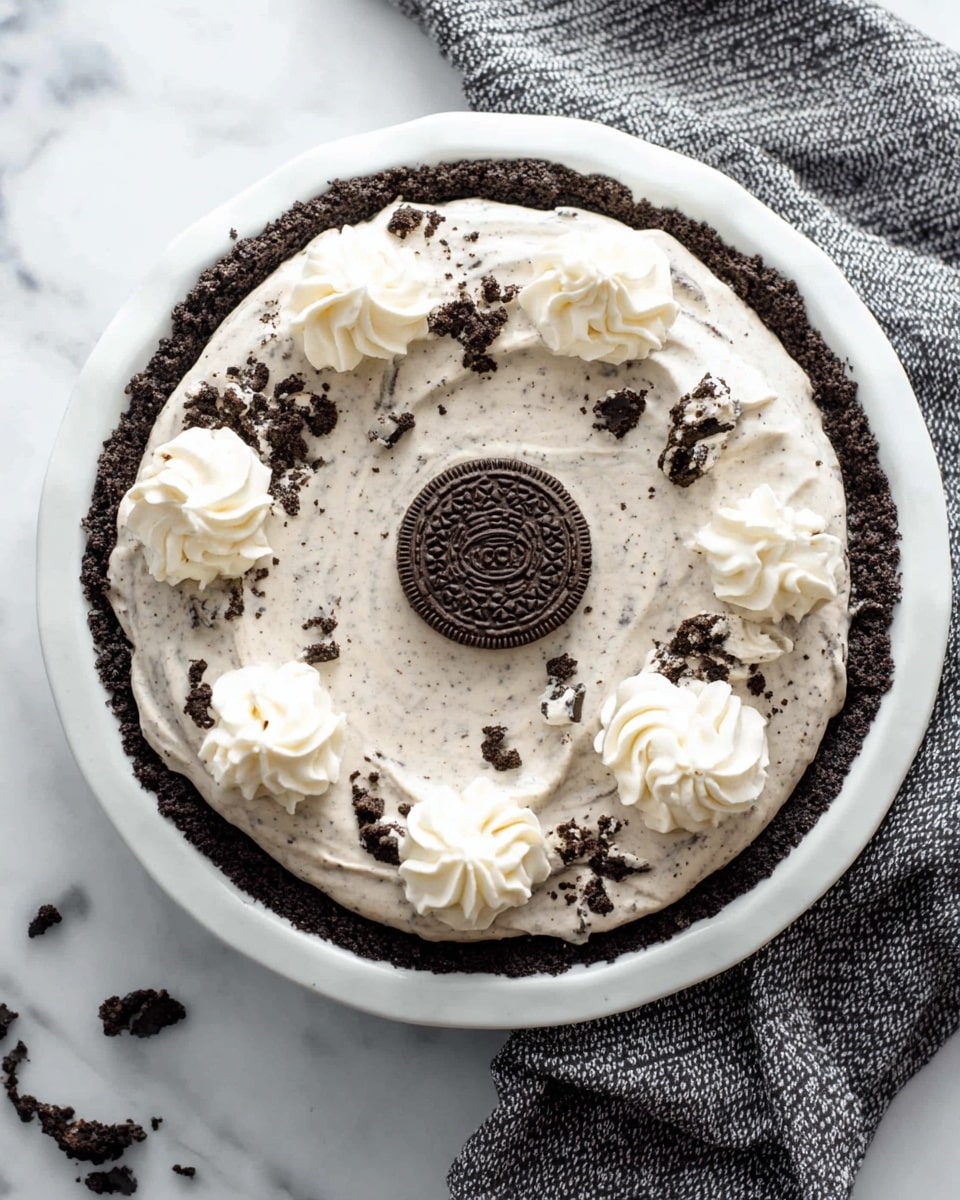 This is a pie with three visible layers. The first layer is a dark chocolate crumb crust that lines the bottom and sides of a white plate. The second layer is a creamy, light gray filling flecked with small dark cookie bits, spread evenly inside the crust. On top of the filling, seven dollops of white whipped cream are spaced around the edge in a circular pattern. Finally, a whole dark chocolate sandwich cookie sits in the center of the pie. The pie is set on a white marbled surface with a textured gray fabric nearby. Photo taken with an iphone --ar 4:5 --v 7