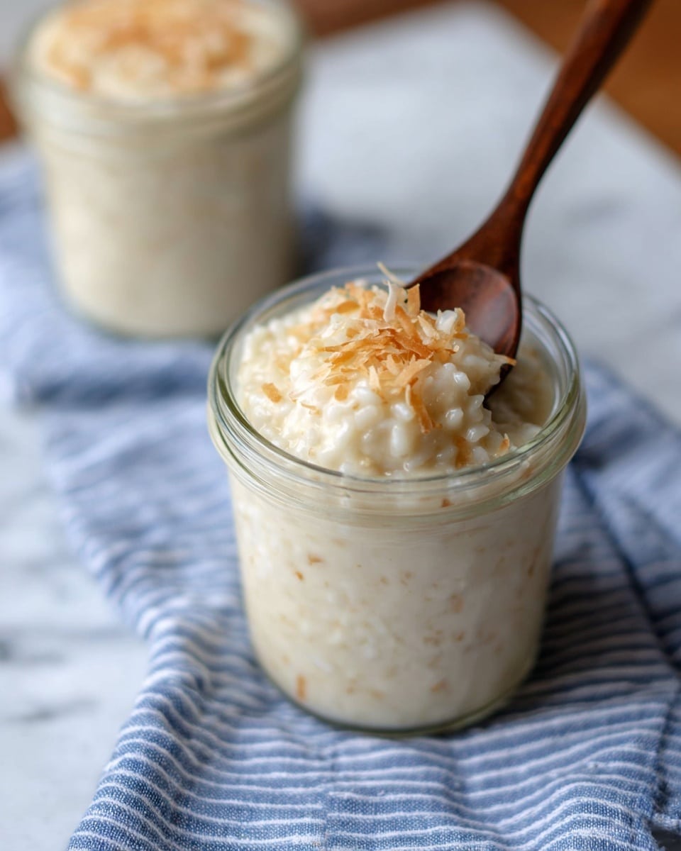 The image shows a creamy, pale beige rice pudding with small visible grains in a clear glass jar. It is topped with a light layer of toasted coconut flakes that add a slightly golden texture and color contrast. A silver spoon is resting on the jar, holding a scoop of the pudding and toasted flakes above the jar. In the background, two more jars of the same pudding are visible on a white, blue-striped cloth over a wooden surface, with an extra spoon lying nearby. The whole scene is set on a white marbled texture. photo taken with an iphone --ar 4:5 --v 7