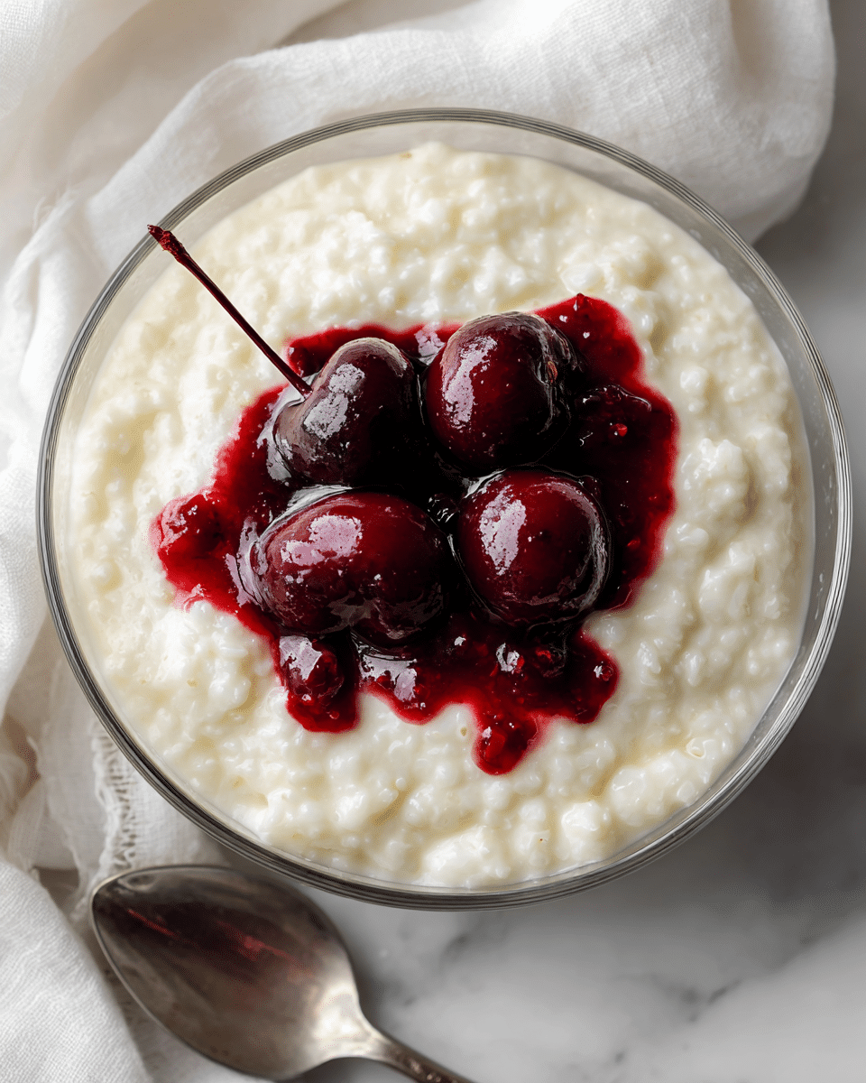 A clear glass bowl filled with a creamy white rice pudding that has a soft and slightly lumpy texture, topped at the center with a small pile of shiny dark red cherries covered in thick cherry sauce that slightly spreads around the cherries. The bowl is placed on a white marbled surface with a blurred white cloth and a silver spoon nearby. photo taken with an iphone --ar 4:5 --v 7