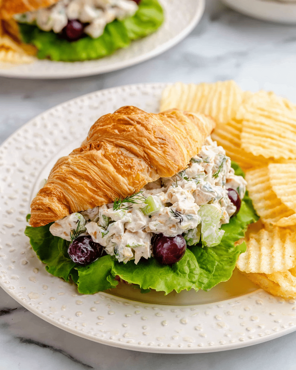 A flaky golden-brown croissant is sliced horizontally and filled with a creamy chicken salad mixed with small chunks of celery, red onion, fresh dill, and halved red grapes. Underneath the chicken salad, there is a crisp green lettuce leaf that adds freshness and color contrast. The croissant sandwich sits on a white plate with raised dotted edges, accompanied by a small pile of ridged, light-yellow potato chips on the right side. The whole scene is set on a white marbled surface, showing another plate with a similar sandwich blurred in the background. Photo taken with an iphone --ar 4:5 --v 7