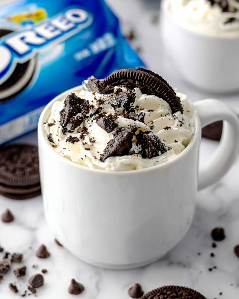 A white mug filled with a creamy white whipped topping forms the base layer, generously covered by broken pieces of dark chocolate Oreo cookies scattered on top. The mug is set on a white marbled surface, surrounded by small chocolate chips and cookie crumbs. In the background, a blue Oreo cookie package is partially visible, creating a bright contrast with the white mug. Nearby, another white mug with a similar dessert can be seen slightly out of focus, adding depth to the scene. photo taken with an iphone --ar 4:5 --v 7