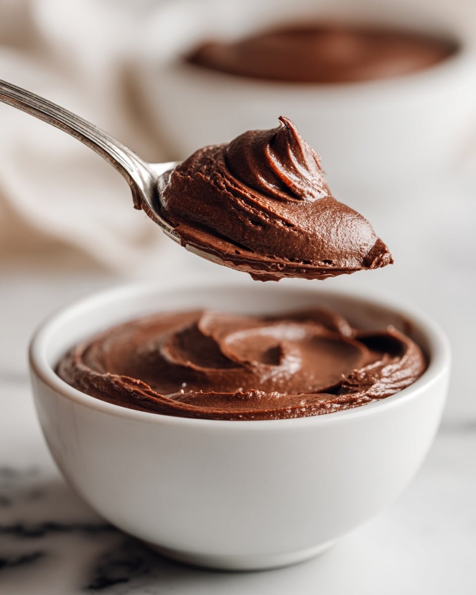 A close-up view of a silver spoon holding a smooth, thick layer of dark brown chocolate mousse, which looks creamy and glossy, lifted above a white bowl filled with more of the same rich mousse. The bowl sits on a white marbled surface, with a blurred background showing a hint of another white bowl with chocolate mousse. Photo taken with an iphone --ar 4:5 --v 7