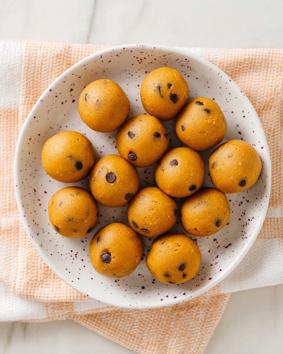 A white plate with an orange and purple speckled pattern holds 15 round, smooth, golden brown cookie dough balls with small dark chocolate chips scattered inside. The balls are arranged randomly but closely packed, showing a soft and slightly cracked texture. The plate rests on a peach and white checkered cloth on a white marbled surface. Photo taken with an iphone --ar 4:5 --v 7