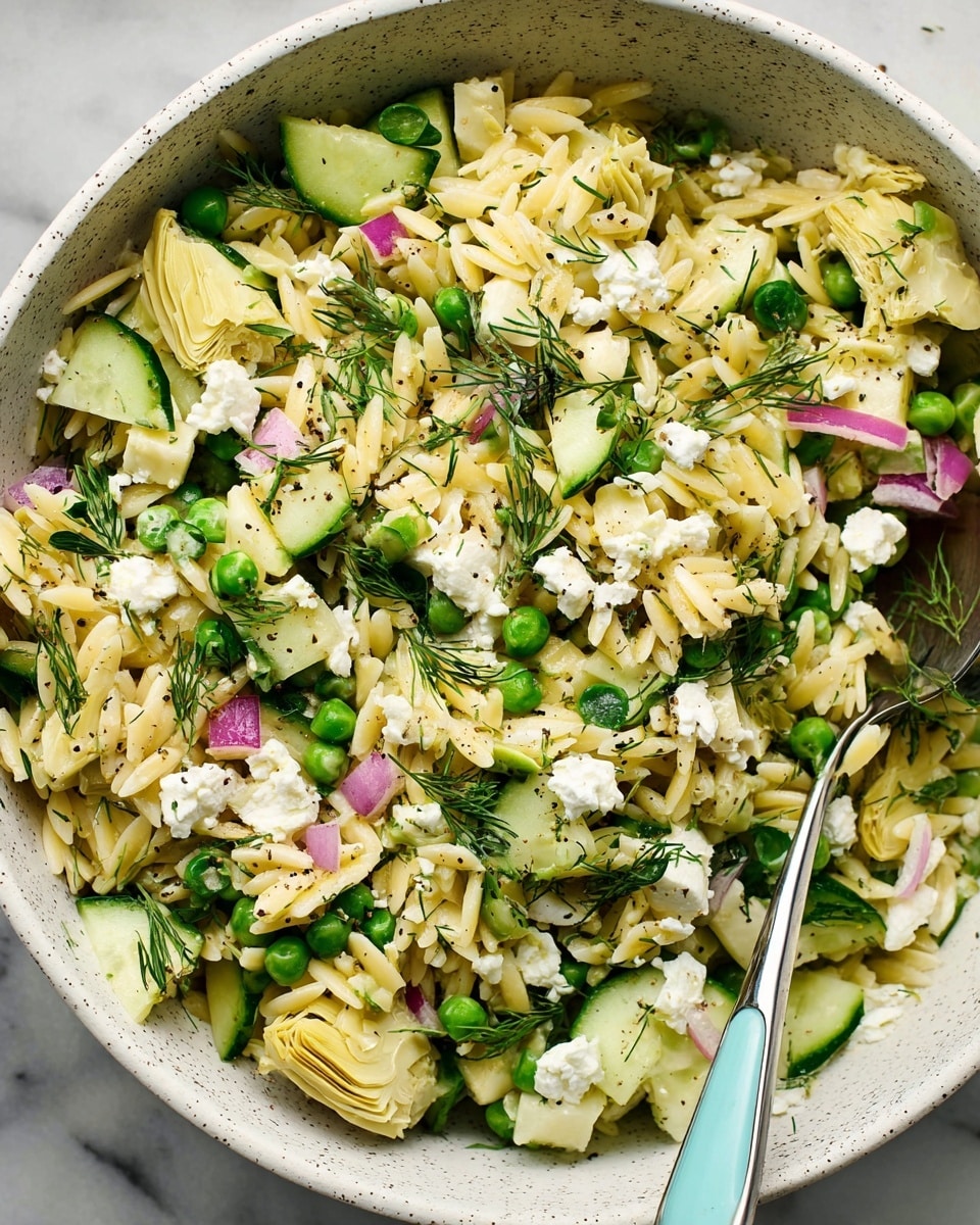 A close-up view of a bowl filled with a layered orzo salad. The bottom layer is light yellow orzo pasta scattered throughout. Mixed on top are chunky pieces of pale yellow artichoke hearts, bright green peas, and small cubes of cucumber with dark green skin. There are small bits of finely chopped red onion and fresh green dill leaves adding color and texture. Crumbled white feta cheese is sprinkled across the salad with some black pepper specks, and a silver spoon with a light blue handle rests inside the bowl. The bowl itself is white with speckles, set on a white marbled surface. photo taken with an iphone --ar 4:5 --v 7