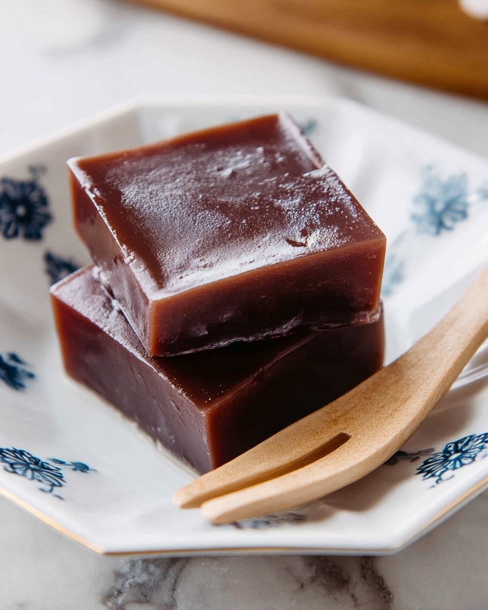 Two square, glossy, dark brown jelly-like pieces stacked on each other are placed on a white octagonal plate with small blue designs along the edges. The top piece shows a smooth, slightly shiny surface with a soft texture, while the bottom piece looks a bit more matte with rounded edges. A light wooden fork rests partially under the top piece on the plate, and the plate sits on a white marbled surface. photo taken with an iphone --ar 4:5 --v 7