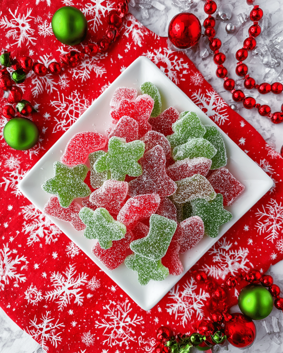 A white square plate filled with two layers of red and green sugar-coated gummy candies shaped like stars, stockings, candy canes, and bells, each candy covered in a fine layer of sugar crystals giving a sparkling texture; the plate rests on a red cloth decorated with white snowflake prints, surrounded by red and white round bead garlands and small green and red decorative berries, all placed on a white marbled texture surface. photo taken with an iphone --ar 4:5 --v 7