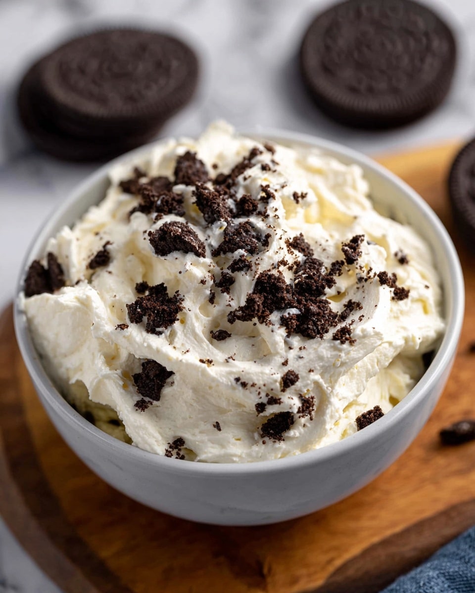 A bowl filled with a creamy white mixture that has a thick and fluffy texture, topped with small dark brown cookie pieces scattered on the surface. The bowl is white, placed on a wooden board, with two whole dark brown cookies visible in the background on a white marbled surface. The cream looks soft and rich, while the cookie crumbs add a rough contrast on top, making the dish look inviting and tasty. Photo taken with an iphone --ar 4:5 --v 7