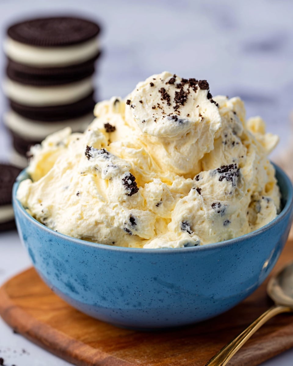 A close-up view of a blue bowl filled with a creamy, fluffy mixture that is pale yellow in color with visible small dark cookie pieces mixed in and sprinkled on top. The texture looks smooth and light, with small chunks adding contrast. The bowl sits on a wooden board placed on a white marbled surface, and in the background, there are three stacked dark chocolate sandwich cookies with white filling, slightly out of focus. photo taken with an iphone --ar 4:5 --v 7