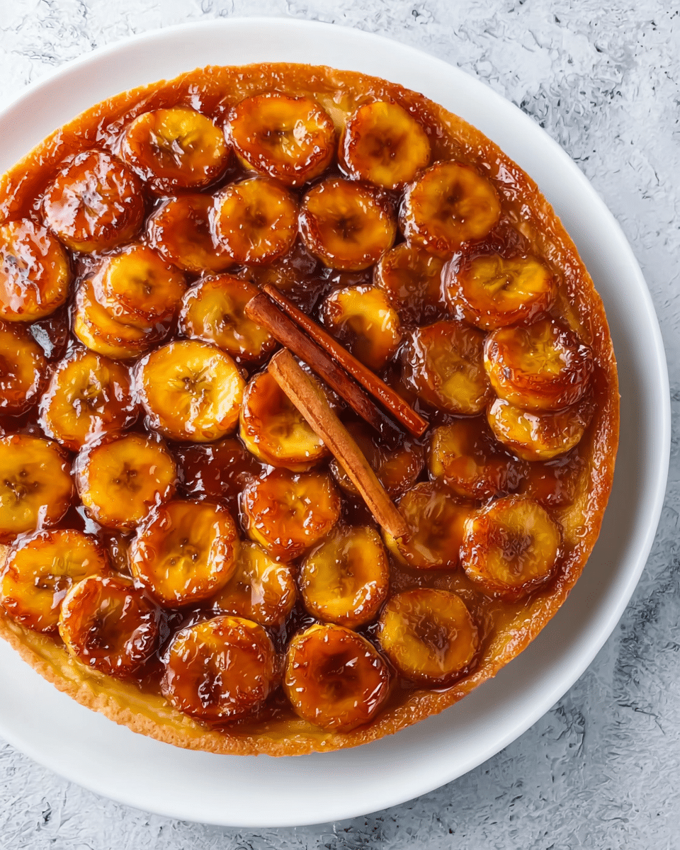 A round tart with a golden-brown crust forms the base layer, holding a glossy caramelized topping of evenly cut banana slices in shades of deep amber and light yellow, arranged closely together covering the entire surface. A single cinnamon stick sits diagonally on top of the bananas, adding texture contrast and warm brown color. The tart sits on a white plate placed on a white marbled textured surface. The caramel sauce glistens over the bananas, giving a shiny and sticky look. photo taken with an iphone --ar 4:5 --v 7