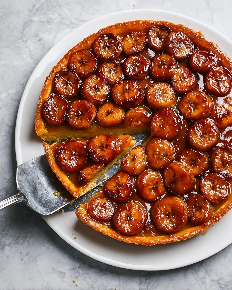 A round tart with three visible layers: the bottom is a golden brown crust that is slightly thick and crisp, the middle is a shiny, caramel amber sauce layer, and the top layer is made up of closely placed round banana slices caramelized to a deep brown with a glossy coating. The tart is on a white plate, and a silver pie server is lifting a slice, showing the crust's texture. The plate sits on a white marbled surface. Photo taken with an iphone --ar 4:5 --v 7