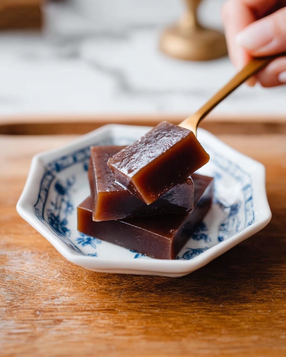 The image shows two square pieces of dark brown jelly-like dessert with a smooth and shiny surface, stacked slightly on a small white octagonal dish with blue floral patterns around the rim. One piece is lying flat, while the other is resting on top at an angle. A woman's hand is holding a small wooden fork, gently touching the top piece, ready to take a bite. The dish is placed on a wooden surface with a white marbled texture in the background, adding a soft and cozy atmosphere. photo taken with an iphone --ar 4:5 --v 7