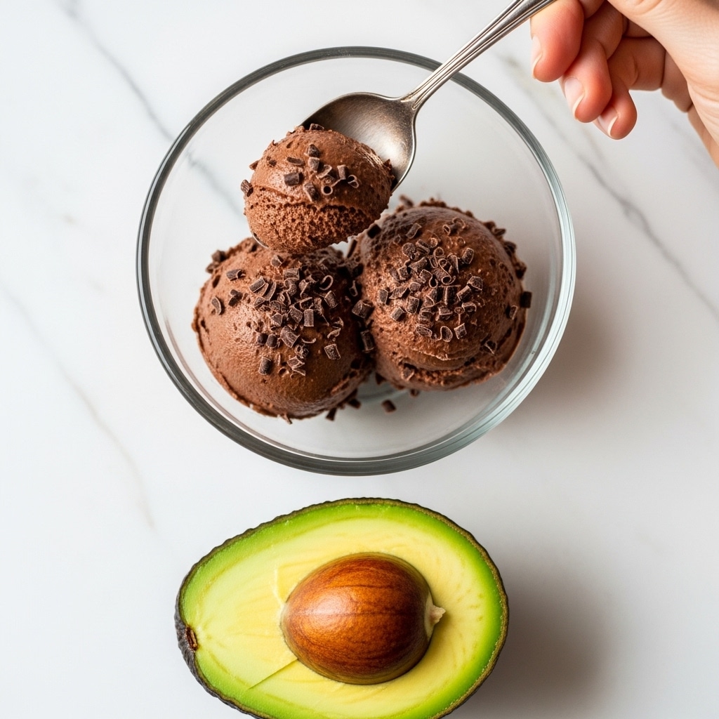 A close-up image shows a clear glass bowl filled with two scoops of dark brown chocolate mousse sprinkled with small chocolate shavings. A vintage silver spoon scoops some of the mousse, held by a woman's hand above the bowl. Below the bowl, there is a fresh avocado cut in half, showing its vibrant light green flesh and a large dark brown pit in the center. The background is a white marbled texture. photo taken with an iphone --ar 4:5 --v 7