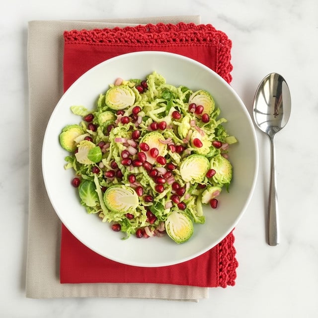 A white shallow bowl filled with a mixed salad of thinly sliced light green Brussels sprouts leaves, scattered with bright red pomegranate seeds and small bits of pink shallots, all tossed together with a light dressing giving a slightly glossy look; the bowl sits on two folded napkins, a larger plain beige one underneath and a smaller red one with a crochet edge on top, placed on a white marbled textured surface. To the right of the bowl is a shiny silver spoon. Photo taken with an iphone --ar 4:5 --v 7
