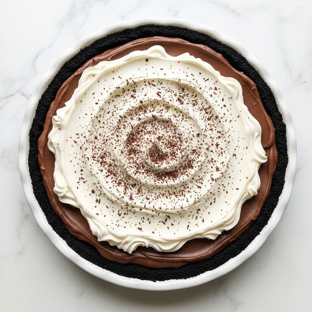 A round dessert with three visible layers sits in a white pie pan on a white marbled surface. The bottom layer is a dark, crumbly chocolate crust that lines the pan completely. Above the crust is a thick, smooth, dark brown chocolate filling. The top layer is a generous swirl of white creamy whipped topping, unevenly spread with soft peaks and valleys. The whipped topping is lightly dusted with finely grated dark chocolate, concentrating more toward the center in a gentle spiral pattern. photo taken with an iphone --ar 4:5 --v 7