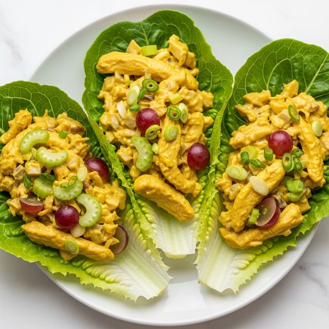 Three lettuce leaves form the base layer, bright green with a fresh, slightly crinkled texture, each leaf shaped like a boat to hold the filling. On top, a thick layer of creamy chicken salad fills the lettuce leaves, consisting of tender yellowish chicken strips mixed with a smooth, golden-yellow sauce. Scattered throughout the chicken salad are light green celery slices and small pieces of red grapes, adding contrasting colors and textures. Thin slices of green onion are sprinkled on top, adding a fresh and sharp touch. All of this is presented on a clean white plate resting on a white marbled surface. Photo taken with an iphone --ar 4:5 --v 7