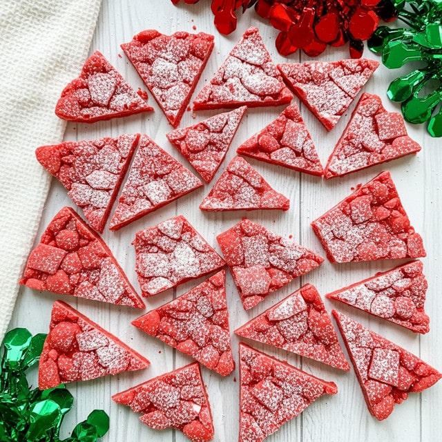 The image shows many pieces of bright red candy bark scattered on a white marbled wood surface. The candy pieces are cut into uneven shards, mostly triangular and square shapes, each covered with a light dusting of white powdered sugar that gives a frosted look. Around the candy, there are sections of shiny green and red foil decorations adding a festive touch, with one white textured cloth partially visible on the left side. The overall feel is bright, colorful, and crunchy, capturing the holiday spirit. photo taken with an iphone --ar 4:5 --v 7
