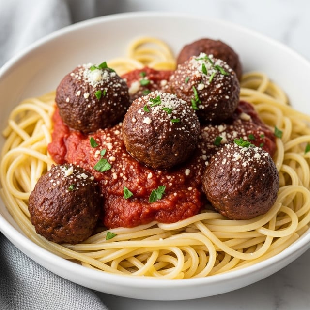 A close-up view of a serving of spaghetti with meatballs in a white bowl. At the bottom, there is a layer of long, light yellow spaghetti noodles covered partially by a thick, deep red tomato sauce. On top of the sauce, five round meatballs of dark brown color are scattered, some with sauce on top and sprinkled with small bits of grated white cheese and green parsley. The bowl rests on a soft grey cloth, all set on a white marbled surface. photo taken with an iphone --ar 4:5 --v 7
