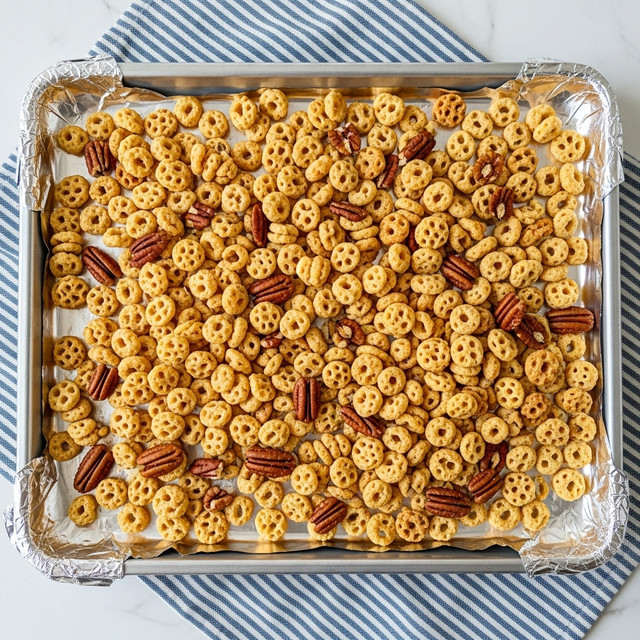 The image shows a baking tray lined with shiny aluminum foil, filled with a mix of small, round, waffle-textured cereal pieces in light yellow and golden brown colors, along with a scattering of pecan halves in dark brown. The tray rests on a blue and white striped cloth on a white marbled surface. The cereal pieces vary slightly in shade and texture, some looking crispier and more toasted than others, giving a crunchy appearance. Photo taken with an iphone --ar 4:5 --v 7