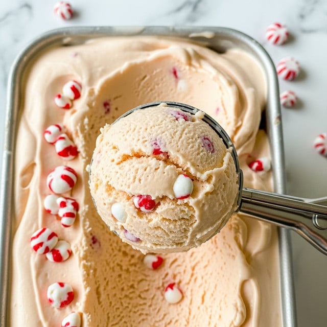 A close-up view of pale peach-colored ice cream with small white and red candy pieces mixed in, showing a creamy and smooth texture. A metal ice cream scoop is holding a rounded, dense scoop of the ice cream in the center, with slightly uneven surface details. The ice cream fills a container with the same soft peach color, and some candy pieces are scattered around the scoop area for texture contrast. The photo's background features a white marbled texture. photo taken with an iphone --ar 4:5 --v 7