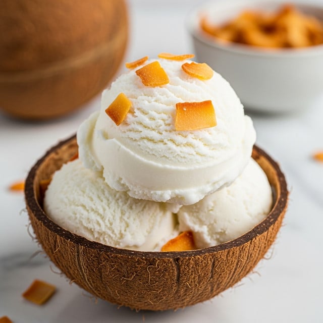 A close-up view of a scoop of white coconut ice cream with a smooth, creamy texture, topped with small pieces of toasted golden-brown coconut flakes, all placed inside a brown, rough-textured coconut shell half, which rests on a white marbled surface with a slightly blurred background showing a second coconut shell half and a bowl with more toasted coconut flakes. photo taken with an iphone --ar 4:5 --v 7