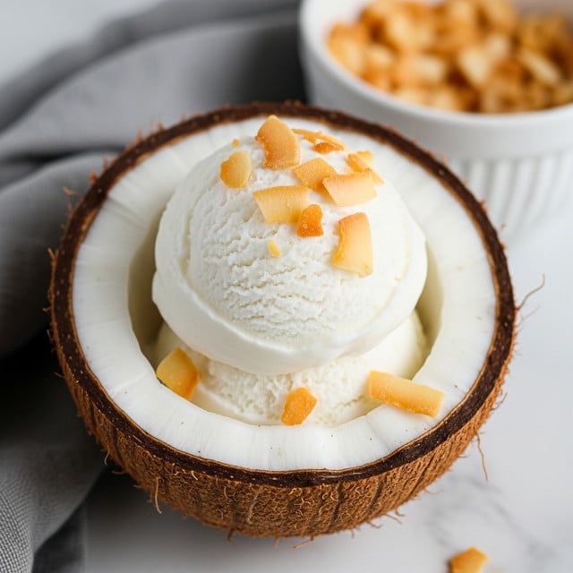 A close-up image shows a halved coconut with its rough brown outer shell and white inner fruit. The hollow coconut half is filled with a smooth, white creamy scoop of coconut ice cream that has a soft, velvety texture. On top of the ice cream, there are small pieces of toasted golden-brown coconut flakes sprinkled evenly. In the background, there is a white bowl filled with more toasted coconut flakes and part of a gray cloth is visible on the white marbled surface. photo taken with an iphone --ar 4:5 --v 7