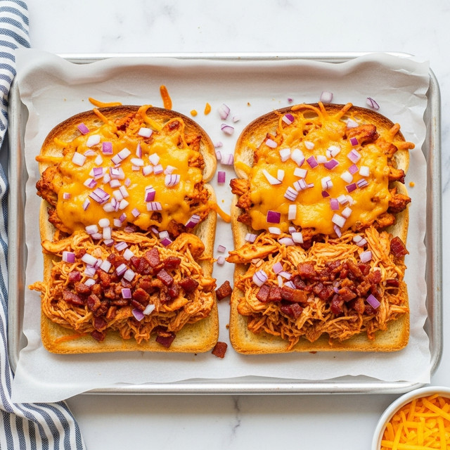Two slices of toasted bread are placed side by side on a baking sheet lined with parchment paper. Each slice has three main layers: the bottom is golden brown toast, the middle is a mix of shredded chicken with small pieces of reddish-brown bacon, and the top is melted cheddar cheese with bits of finely chopped red onion scattered over it. The cheese is melted and slightly browned in some spots, creating a crispy texture. The background is a white marbled texture, with a striped blue and white cloth on the left edge and a small white bowl filled with shredded cheese on the bottom right corner. Photo taken with an iphone --ar 4:5 --v 7