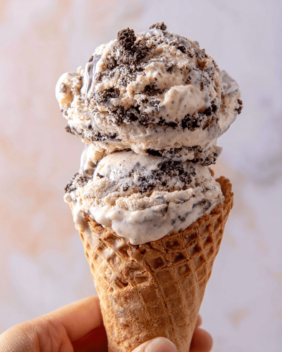A close-up of two scoops of cookies and cream ice cream stacked on top of each other in a light brown waffle cone with a textured grid pattern. The top scoop has a mix of creamy white and dark chocolate cookie pieces, with a slightly melted, creamy texture. The lower scoop also shows the cream and cookie swirls, melting slightly over the cone's edge. A woman's hand holds the cone gently from the bottom left against a soft, blurred background with light pastel tones on a white marbled surface. photo taken with an iphone --ar 4:5 --v 7