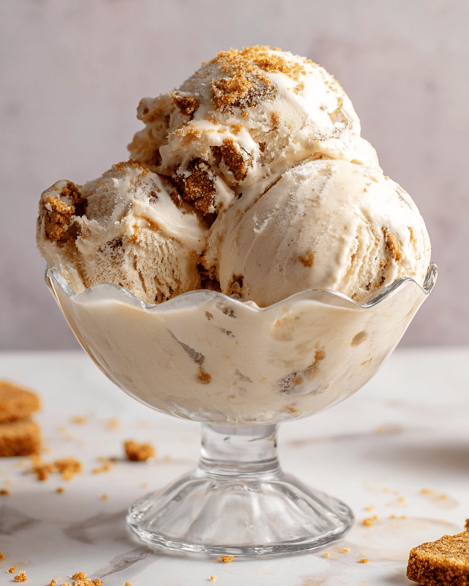 A clear, scalloped glass bowl holds three large scoops of light beige ice cream with visible chunks of brown cookie or biscuit mixed throughout, some melting slightly on the edges. The ice cream has a creamy texture and the cookie bits add a rough, crunchy contrast. There are crumbs scattered around the bowl on a white marbled surface, with a soft, neutral background behind it. Photo taken with an iphone --ar 4:5 --v 7