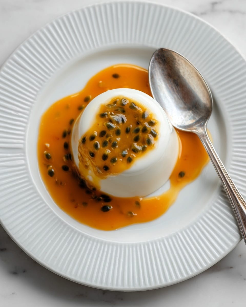 A smooth, white panna cotta dome sits in the center of a white plate with a ridged border, topped with bright orange passion fruit pulp speckled with small black seeds that glisten with moisture. Surrounding the panna cotta is a shiny, amber-colored sauce spread in a loose circular pattern, adding a rich contrast to the dessert. To the right of the panna cotta, a reflective silver spoon rests on the plate, catching soft light. The whole scene is set on a white marbled surface. photo taken with an iphone --ar 4:5 --v 7