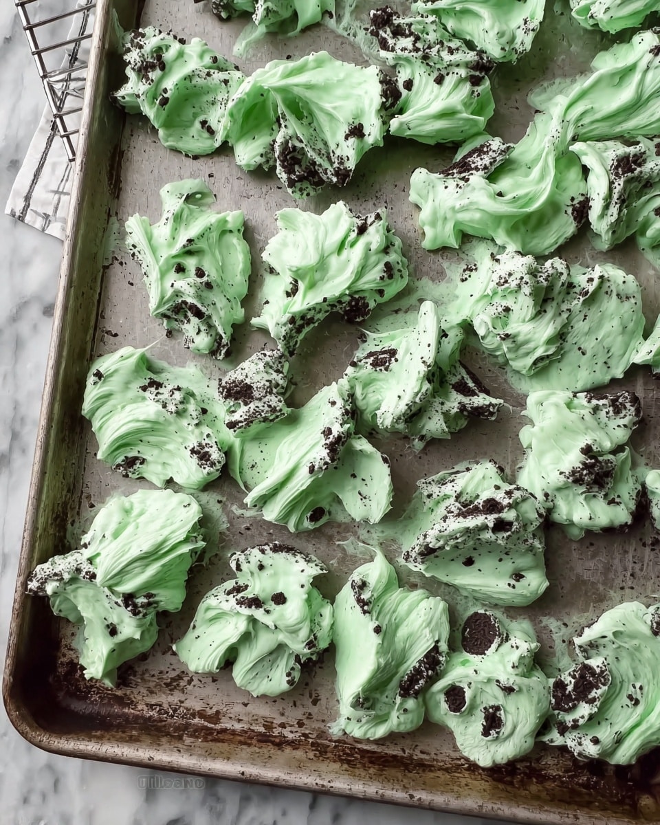 The image shows many small clusters of mint green creamy bark spread unevenly on a used metal baking tray. The bark has a smooth, swirled texture with dark bits of crumbled cookies mixed and visible throughout each cluster, giving a spotted effect. The clusters vary in shape and size, with slightly raised edges and soft, creamy peaks. The tray is placed on a white marbled surface, and part of a cooling rack edge with dark cookies can be seen on the side. Photo taken with an iphone --ar 4:5 --v 7