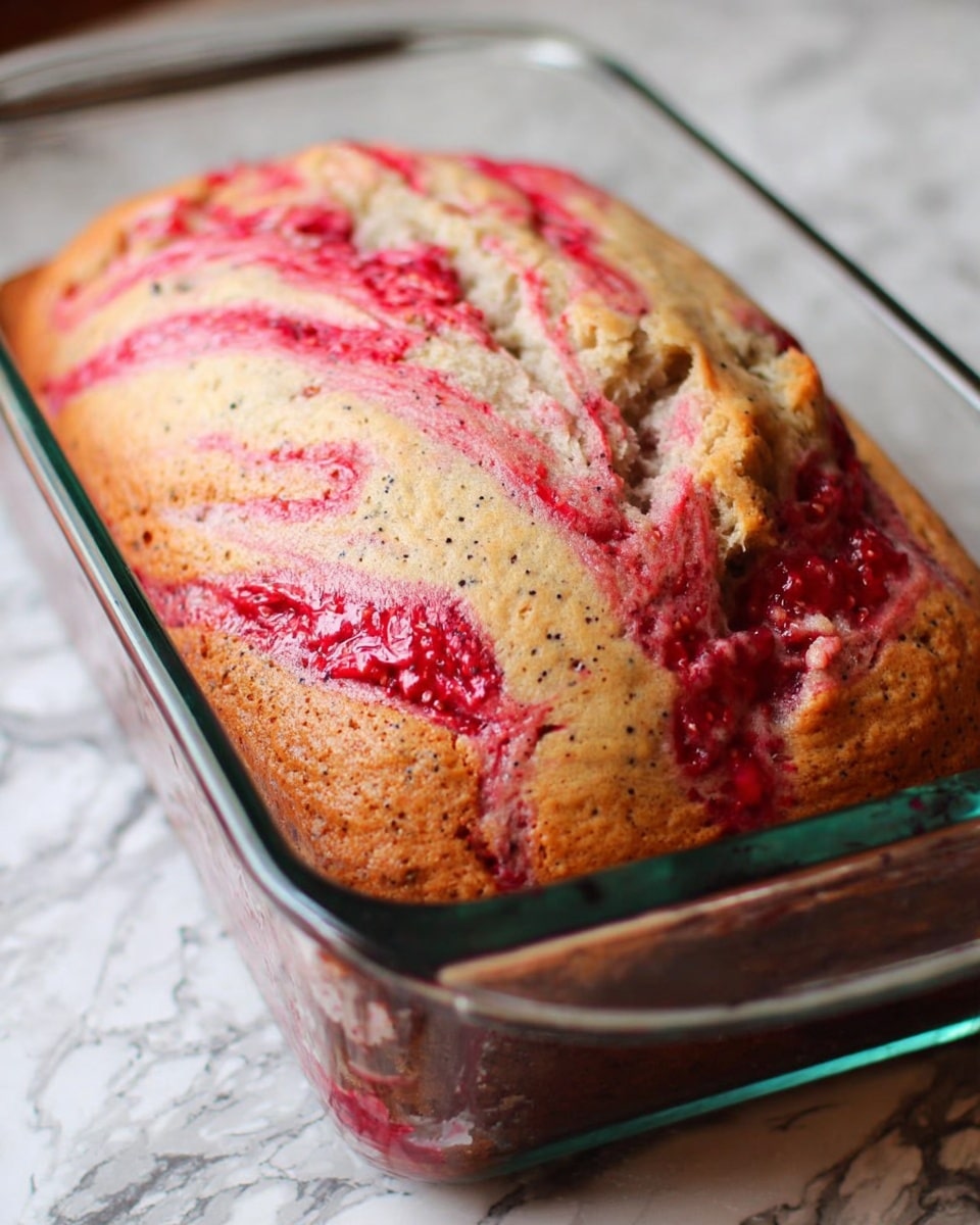 A loaf cake with a golden brown crust and visible small black seeds throughout, topped with bright red swirls of raspberry jam mixed into the light beige batter, baked in a clear glass loaf pan. The texture looks soft and moist with uneven swirled patterns of red and beige on the surface. The loaf pan is placed on a surface with a white marbled texture in the background. photo taken with an iphone --ar 4:5 --v 7