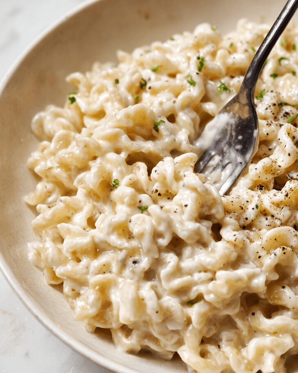 A close-up image of creamy pasta made with short, wavy noodles coated evenly in a thick white sauce. The sauce looks smooth with small specks of black pepper and tiny green herb pieces scattered throughout. A silver fork is partially visible stirring the pasta, with some sauce clinging to its tines. The pasta rests in a white bowl against a white marbled texture background. The overall look is rich and comforting, highlighting the texture of the creamy sauce mixed with the curly pasta shapes. photo taken with an iphone --ar 4:5 --v 7