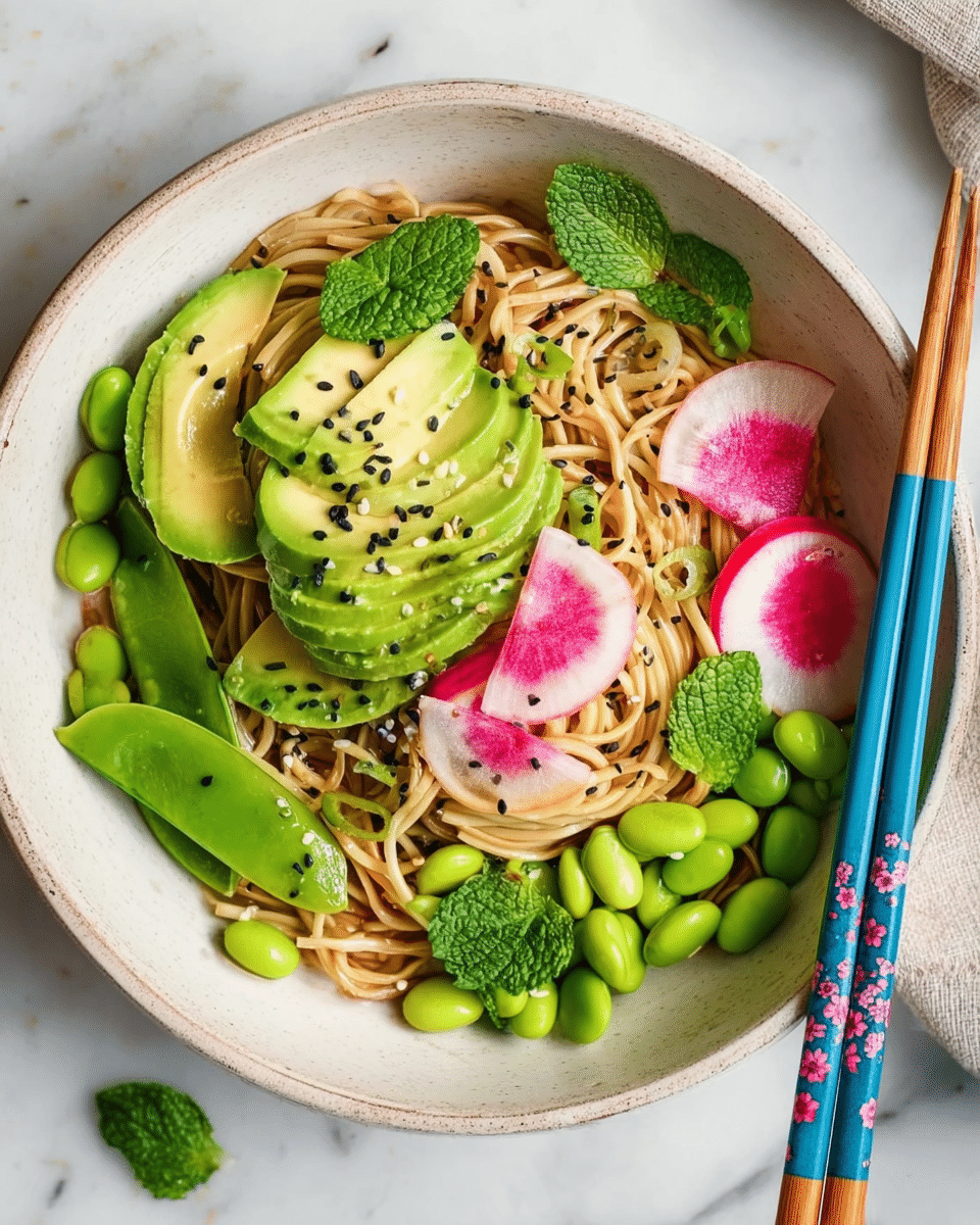 A white bowl on a white marbled surface holds a vibrant noodle dish with several layers, starting with a base of light brown noodles piled softly in the center. On top and around the noodles, bright green sliced avocado fans out on one side, while smooth green edamame beans are scattered throughout. Thin, round slices of pink and white radish are placed on one edge, along with vibrant green snap peas. Fresh mint leaves in deep green are spread evenly across for a touch of freshness. The dish is sprinkled with small black and white sesame seeds, adding texture and detail to the colors. A pair of blue chopsticks with a floral pattern rests on the bowl's edge. photo taken with an iphone --ar 4:5 --v 7