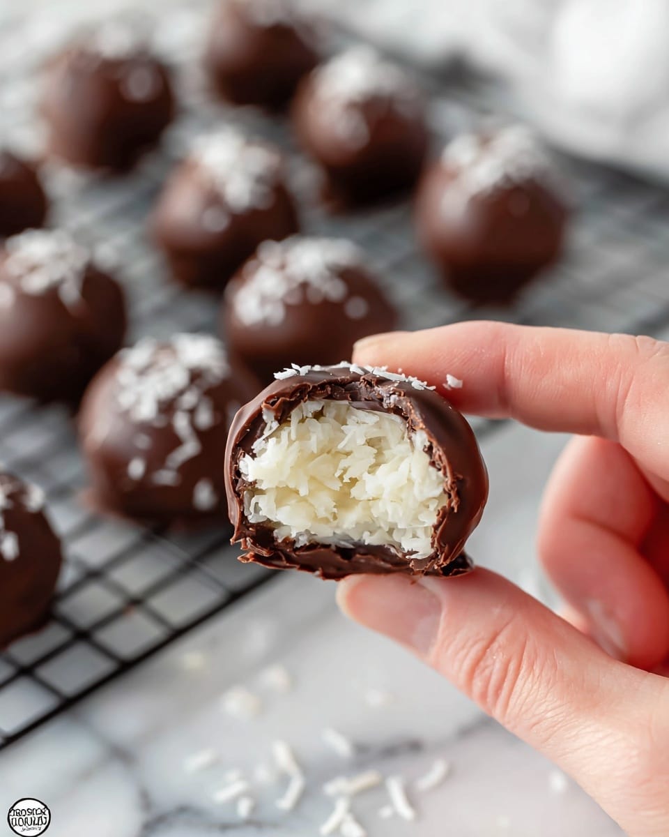A close-up shows a woman's hand holding a small chocolate ball with a bite taken out, revealing a white, creamy, shredded coconut filling inside. The outer layer is a smooth, dark brown chocolate coating that is cracked where the bite is. In the blurred background, several more chocolate balls are placed on a white marbled surface with a wire cooling rack underneath, some sprinkled lightly with white coconut flakes. The overall look is soft and rich. photo taken with an iphone --ar 4:5 --v 7