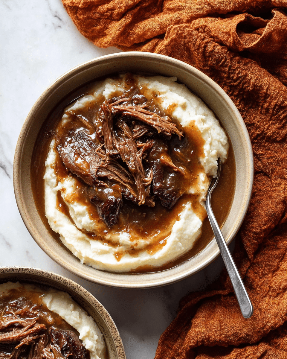 A beige bowl holds a two-layer dish placed on a white marbled surface; the bottom layer is creamy, smooth white mashed potatoes spread evenly across the bowl, topped with several thick pieces of dark brown shredded meat covered in a glossy, rich brown gravy that glistens under the light. A metal spoon rests slightly buried in the mashed potatoes at the front of the bowl. To the right of the bowl, a crumpled orange-brown cloth adds a cozy touch, and part of a second similar bowl is visible nearby. photo taken with an iphone --ar 4:5 --v 7